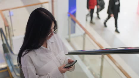 confident woman wearing glasses and white shirt turns while holding phone on escalator in brightly lit mall interior with people in motion and retail background in view