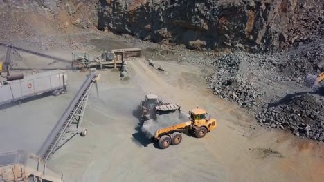 a backhoe excavator pours ground up rock into a dump truck in a quarry next to some conveyor belts