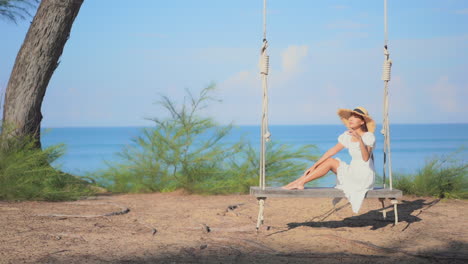 hermosa mujer asiática sentada suavemente en el gran columpio de banco de madera con vestido de verano y sombrero en el fondo azul del mar en tailandia, cámara lenta