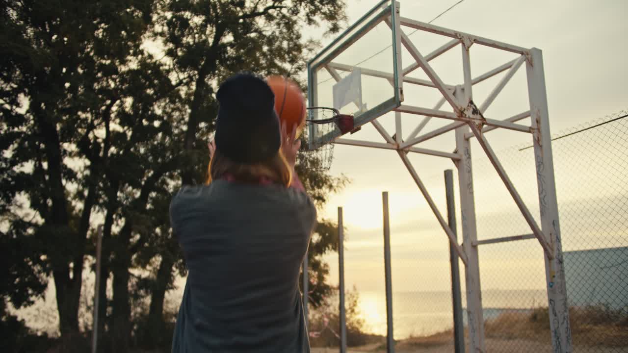 una chica rubia con un sombrero negro y una camiseta gris lanza una pelota de baloncesto naranja al aro durante su entrenamiento de baloncestro y está muy feliz por ello temprano en la mañana al amanecer