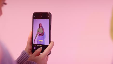 Studioaufnahme-Einer-Frau,-Die-Ein-Foto-Einer-Freundin-Macht,-Die-Auf-Dem-Mobiltelefon-Vor-Rosa-Hintergrund-Tanzt