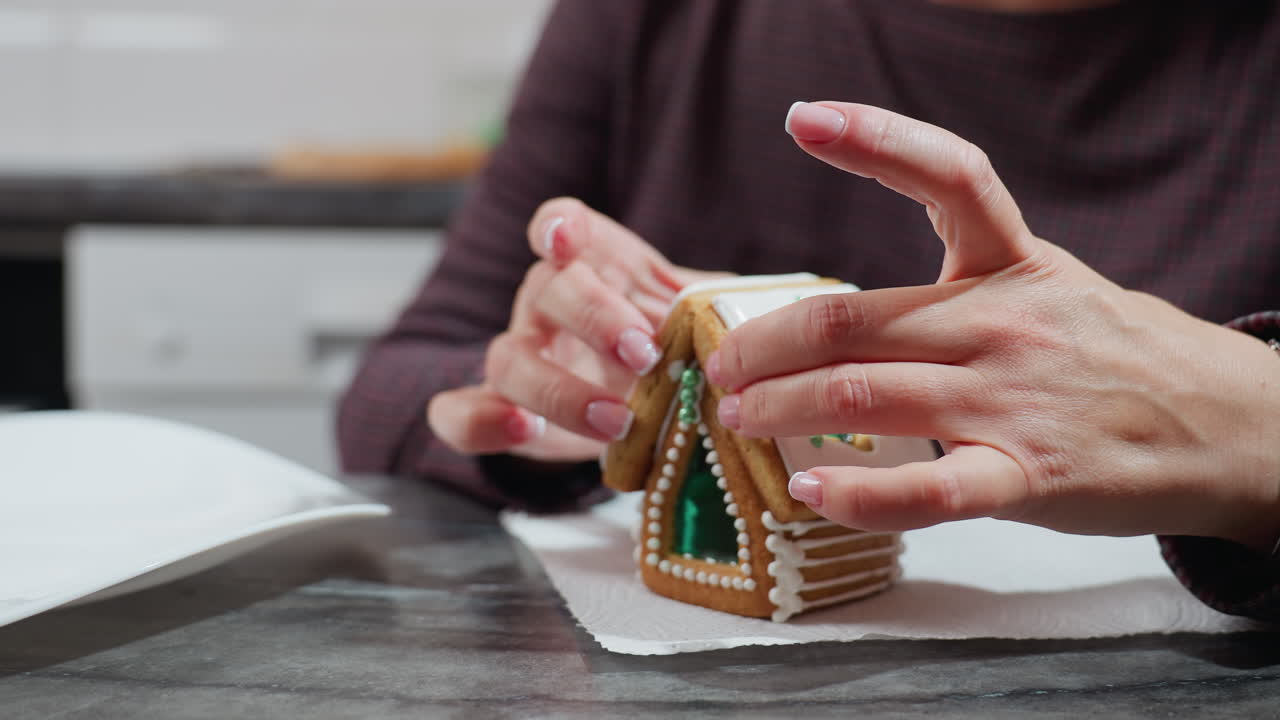 신선한 아이싱과 복잡한 장식으로 새로 조립된 진저브레드 하우스 쿠키를 안정시키는 여성의 클로즈업.