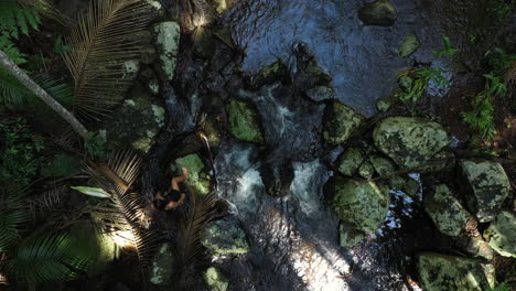 Top-view-drone-shot-of-a-girl-woman-walking-on-rocks-in-a-river-stream-in-a-dense-tropical-forest-woods-in-Australia