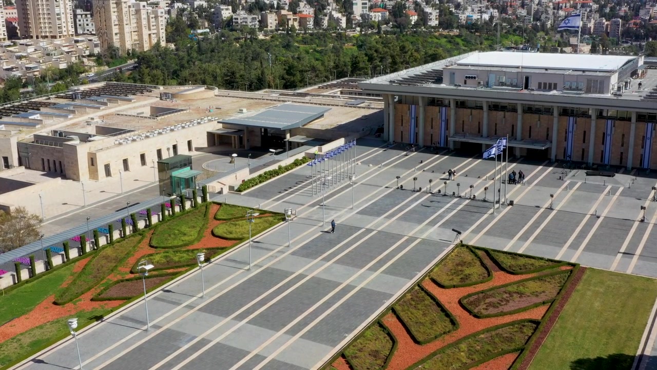 Perspectiva de ângulo alto da fachada do parlamento israelense, com pátio pavimentado e caminhos de jardim bem cuidados, durante um dia ensolarado em Jerusalém.