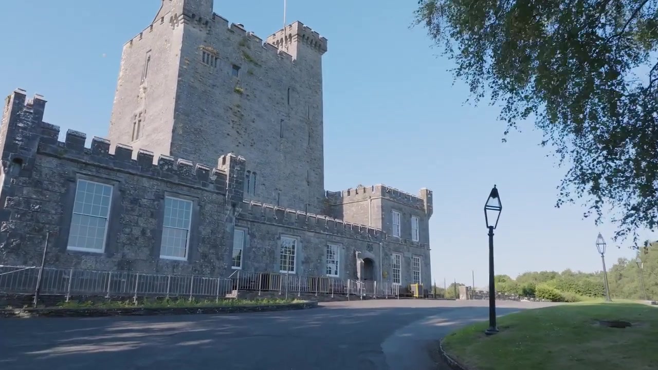 La telecamera scivola lungo un viale asfaltato curvo verso la facciata in pietra del Castello di Knappogue incorniciato da alberi illuminati dal sole