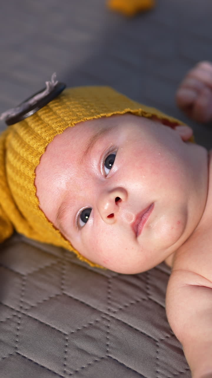 Bare baby in a funny yellow cap lying on the bed. Small boy lying calmly on the bed and look at the camera. Vertical video