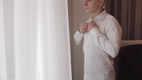 Stylish-man-in-jacket-fastens-buttons,-buttoning-white-shirt-preparing-to-go-out-near-window