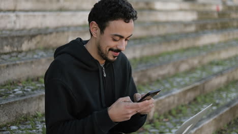 young arabic man with dark curly hair and beard in black hoodie sitting on stairs outside