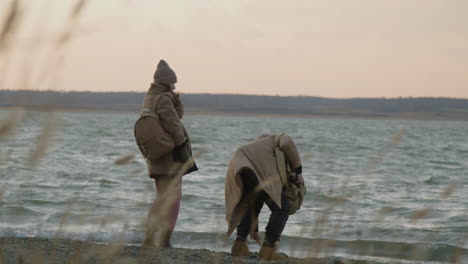 rückansicht von zwei freunden in winterkleidung, die an einem bewölkten tag an einer meeresküste kieselsteine ins wasser werfen 1