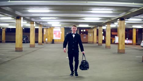 young blonde businessman in a black suit with bow-tie walking on the parking with big leather bag. slow motion