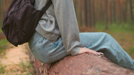 two hikers rest on fallen tree in lush forest, with one hand resting on tree trunk showcasing long nails, and companion in green shirt relaxed nearby, surrounded by tall trees and nature s serenity