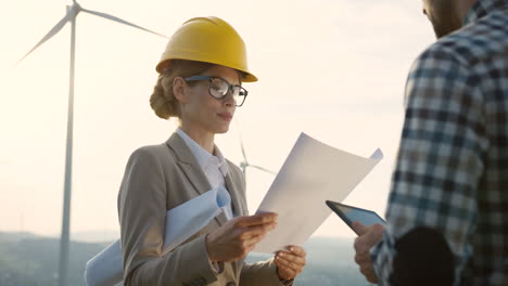 caucasian man and woman engineers wearing a helmet watching some blueprints and using tablet while talking at wind station of renewable energy