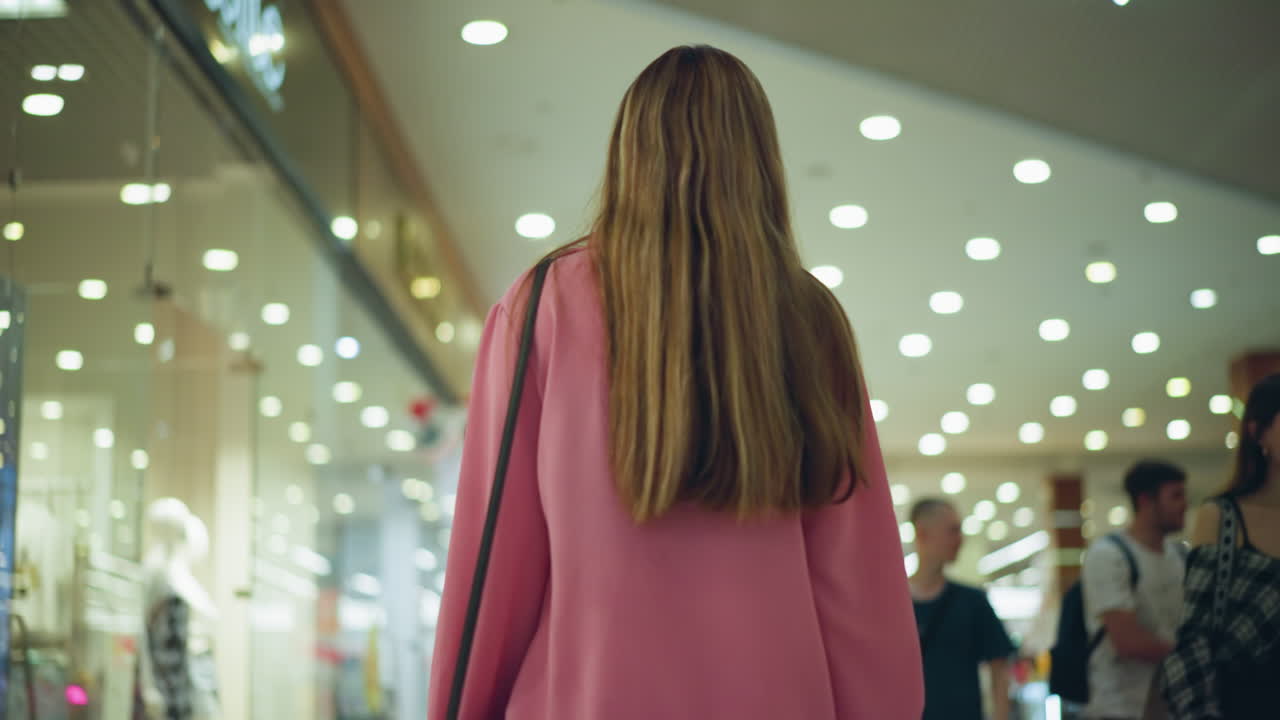Lady in pink dress carrying black bag is seen from behind, walking through a brightly lit mall while looking around, other shoppers walk in opposite directions, with slightly blur view of other items