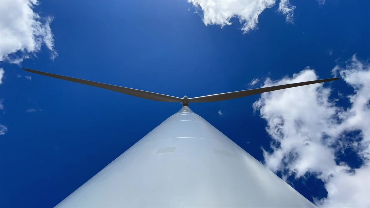 Timelapse che mostra una turbina eolica vista dal basso con pale che si muovono lentamente e nuvole che si spostano, evidenziando l'infrastruttura di energia rinnovabile.