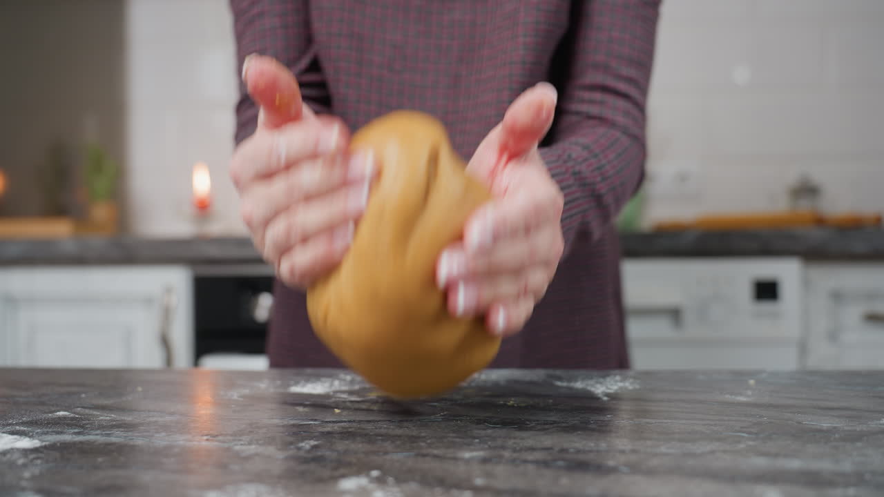 близкий взгляд на женщину, которая мяшает тесто, поднимает его и бросает на кухонный стол, руки в движении, мучная пыль на поверхности, мягкое тесто отскакивает, теплая кухонная обстановка с современными шкафами на заднем плане