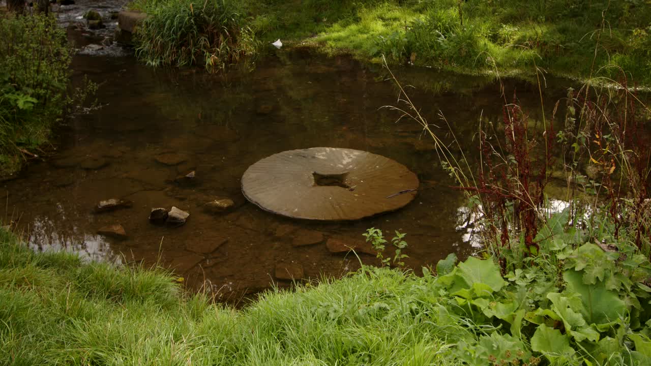 mid shot of disused water mill stone on the river dove with Viator's Bridge in the back ground, Milldale