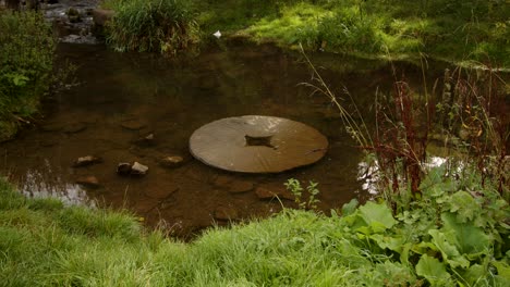 mid-shot-of-disused-water-mill-stone-on-the-river-dove-with-Viator's-Bridge-in-the-back-ground,-Milldale