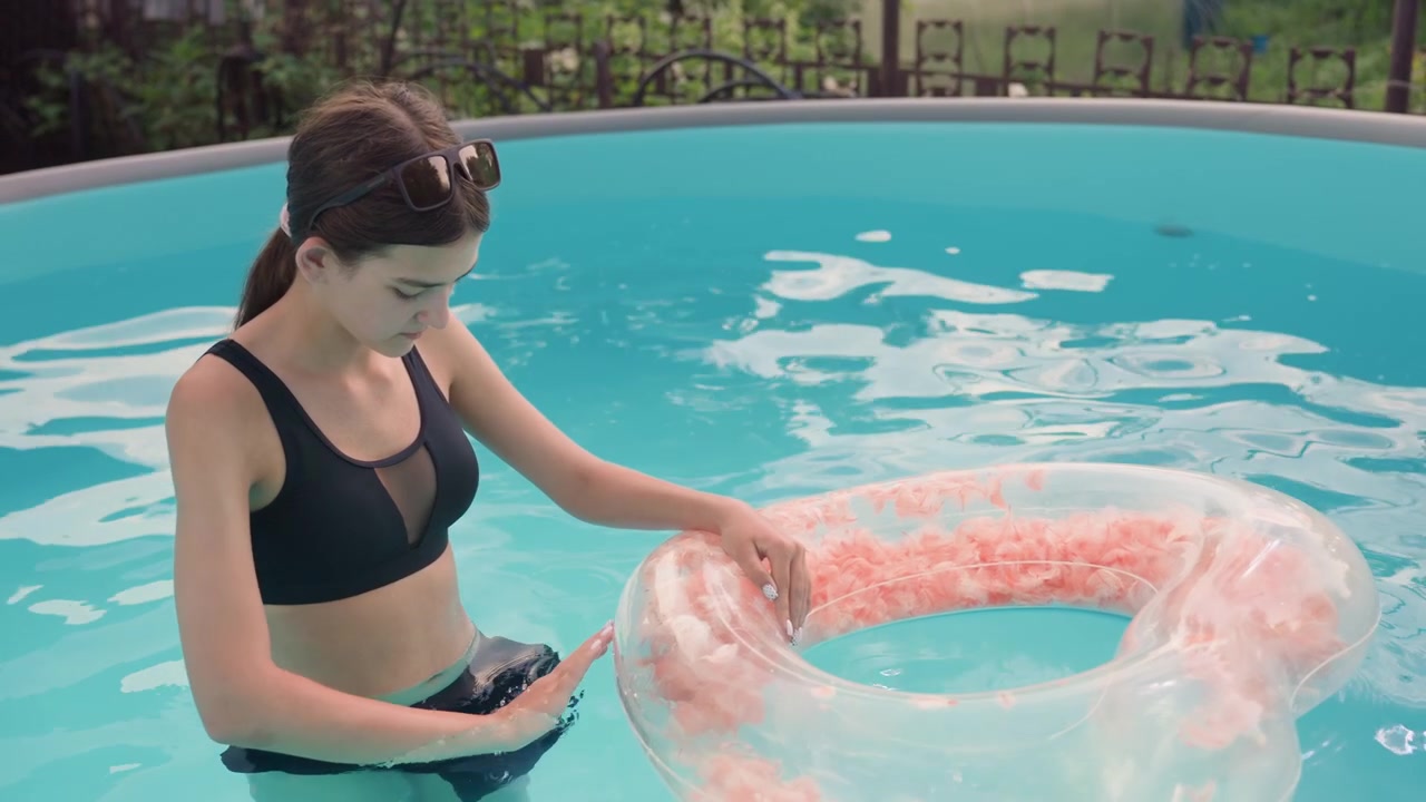 Young woman in black swimsuit interacts with inflatable pool ring while standing waist deep in water. Woman touches clear tube which contains pink material inside as woman remains still