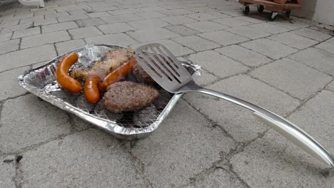 locked off view of disposable mini bbq grill with different meats and spatula leaning against it