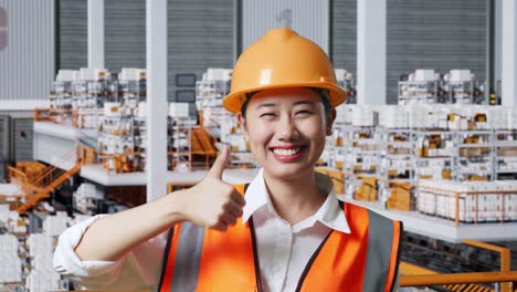 close up of asian female engineer with safety helmet standing in the warehouse with shelves full of delivery goods. smiling and showing thumbs up gesture to the camera in the storage