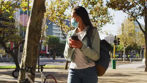 Mujer-Asiática-Con-Mascarilla-Usando-Un-Teléfono-Inteligente-De-Pie-En-El-Parque