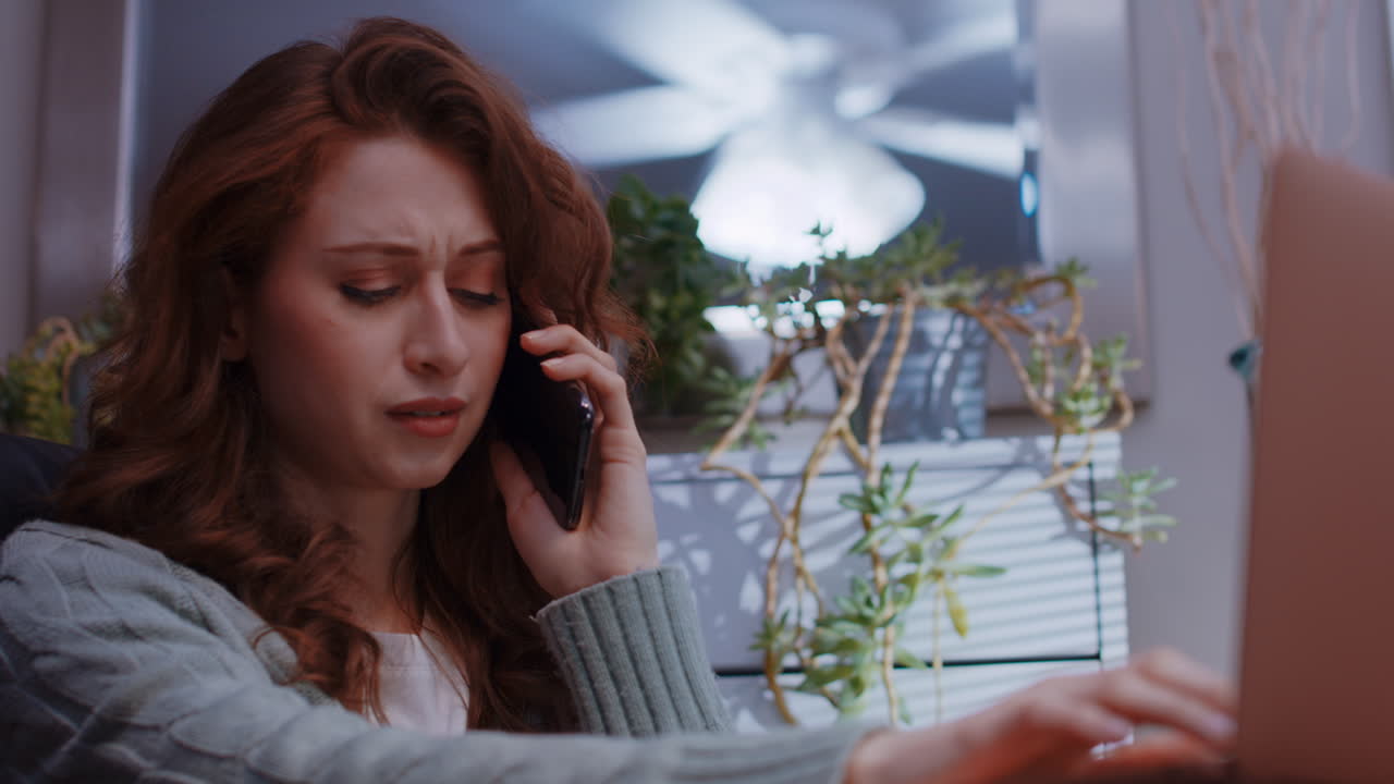 una joven mujer caucásica está trabajando desde casa en su computadora portátil, recibe noticias tristes por teléfono