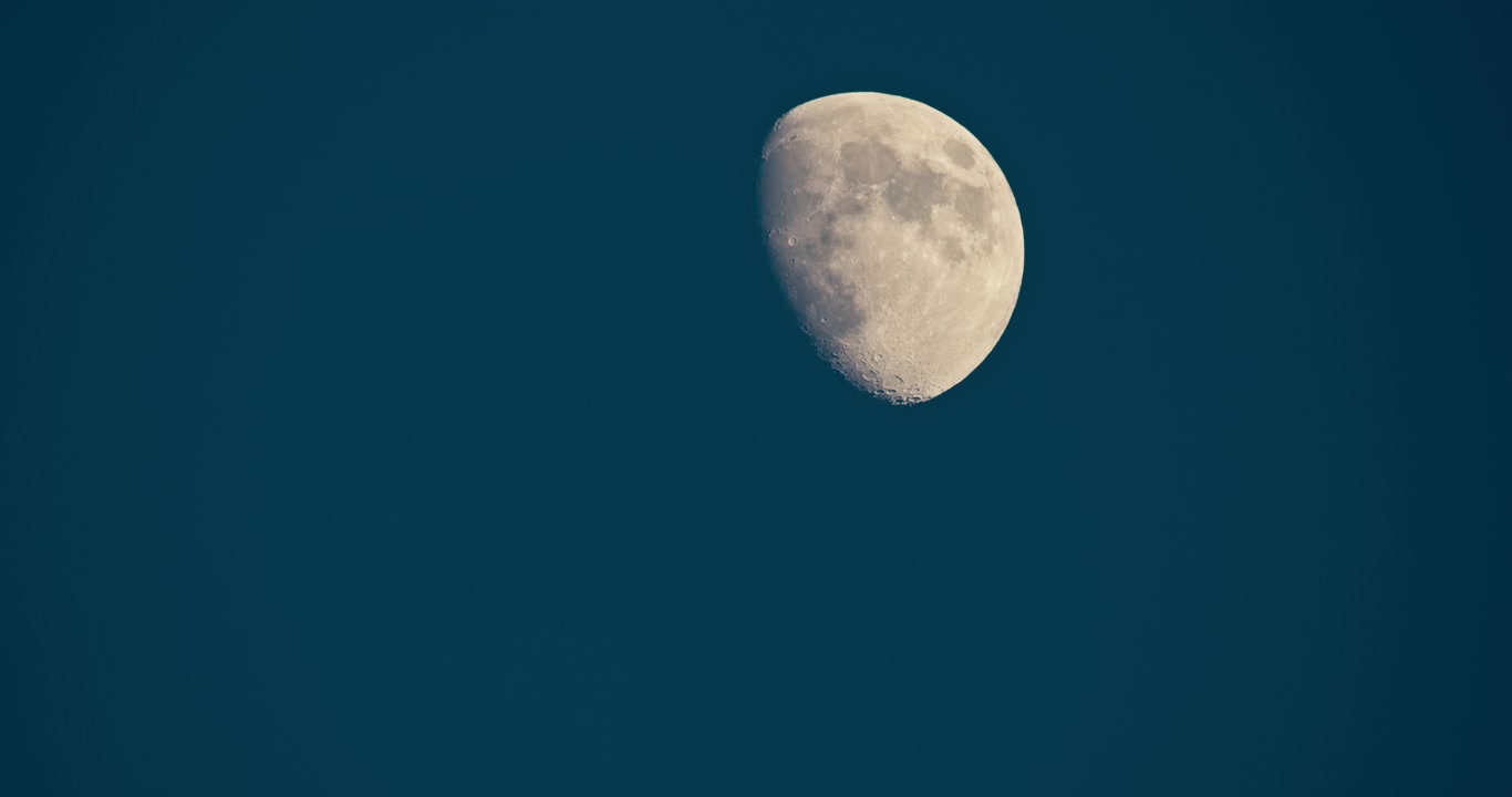 Vista em close-up de uma lua gibosa crescente contra um céu noturno azul profundo. Superfície lunar detalhada com uma composição limpa e minimalista e amplo espaço para texto.