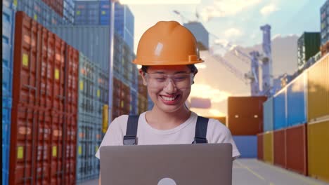close up de una trabajadora asiática que usa gafas de protección y casco de seguridad sonriendo y señalando una computadora portátil en su mano mientras está de pie en el almacén del patio de contenedores