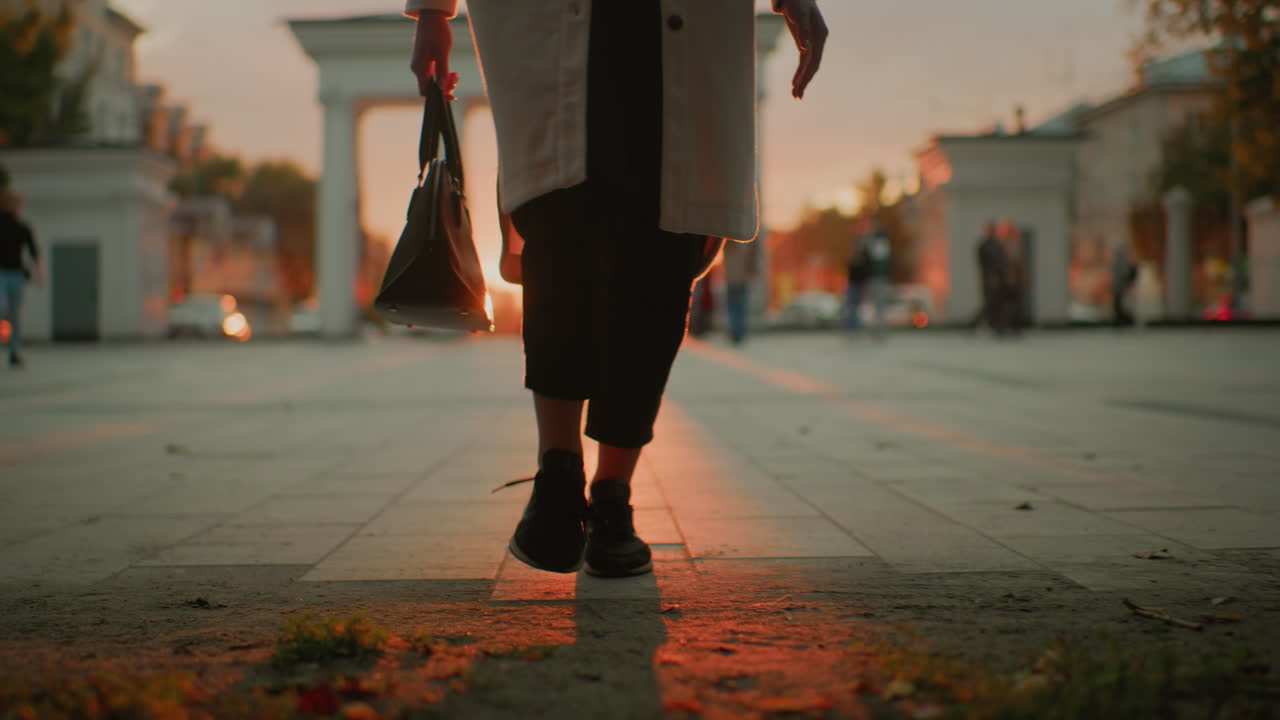 lower body view of girl in long white coat strolling with stylish handbag while golden sunlight creates warm glowing effect during sunset in vibrant urban environment with people and cars moving