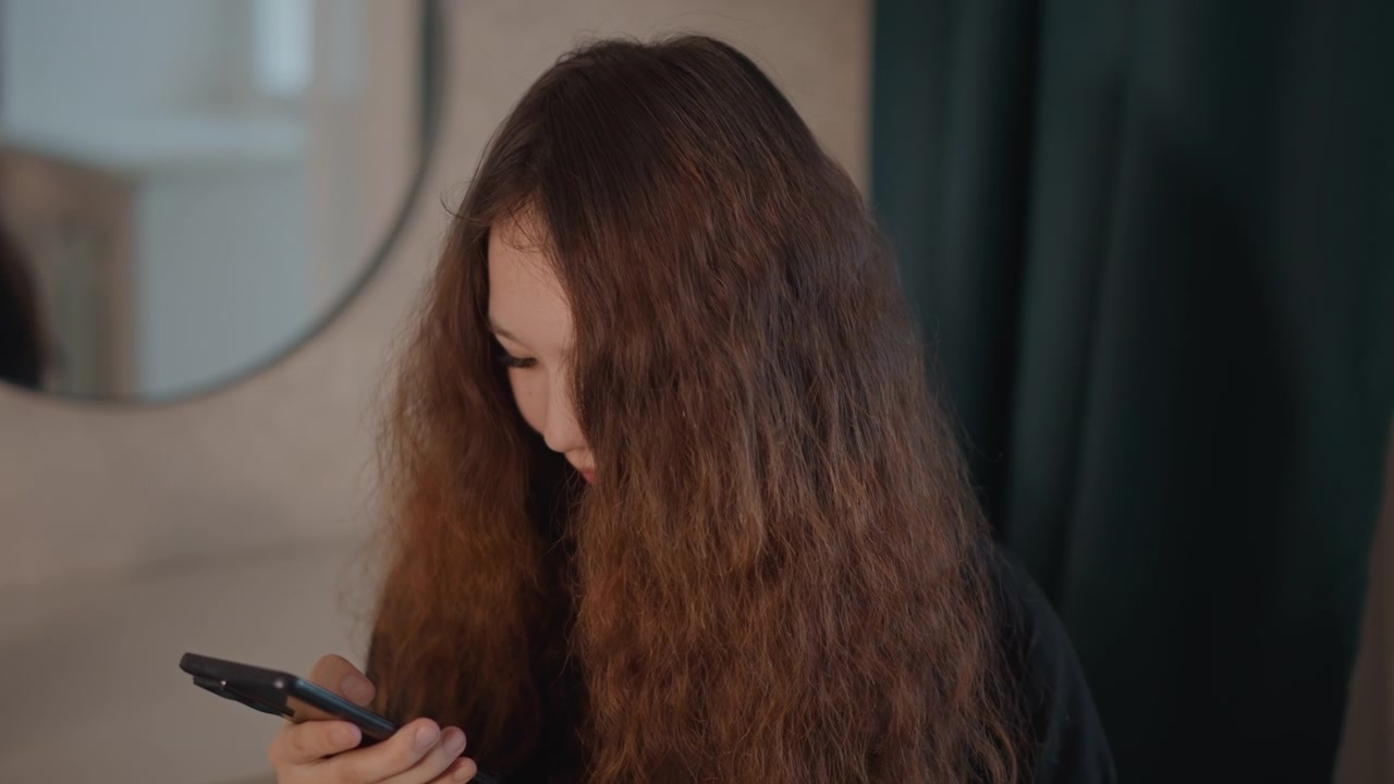 Mujer joven sentada en interiores usando un smartphone con suaves gestos de desplazamiento y toque, cabeza ligeramente inclinada, hombros relajados, luz cálida y difusa, en un momento privado y tranquilo y un entorno hogareño natural.