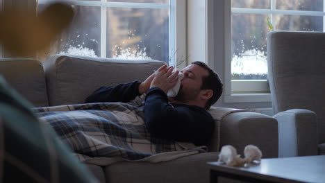 man with flu sneezing into napkin while lying down on sofa in living room