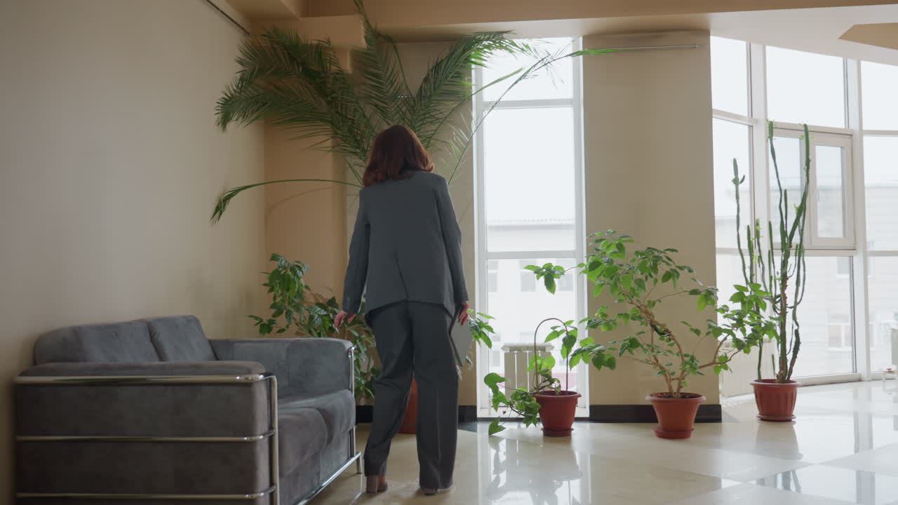 Businesswoman in gray suit walks confidently through bright office lobby filled with potted plants and natural light, heading toward seating area with calm demeanor and professional posture