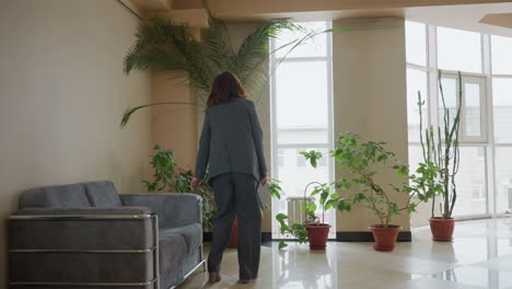 businesswoman in gray suit walks confidently through bright office lobby filled with potted plants and natural light, heading toward seating area with calm demeanor and professional posture