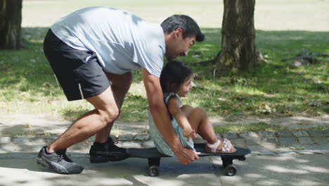 side view of happy asian man giving his daughter ride on skateboard in park