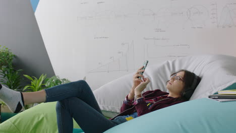 happy woman student using smartphone browsing online social media messages enjoying lunch break relaxing on colorful pillows listening to music in modern office