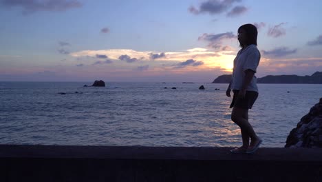 Follow-tracking-shot-girl-in-summer-outfit-walking-on-sea-wall-next-to-ocean-at-sunset