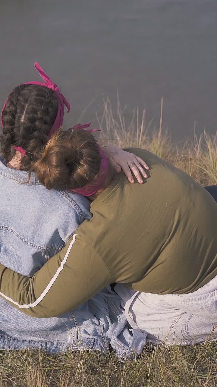 young women hug having date on steep bank of calm river in warm autumn evening close backside view
