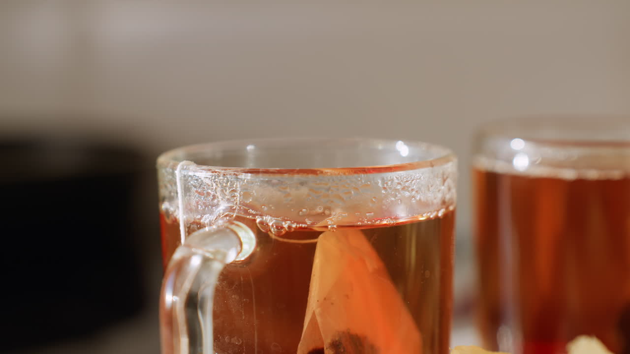 Primo piano di tè caldo fumante in tazza di vetro con bustina all'interno, condensa che si forma sul bordo e liquido ambrato caldo che brilla alla luce del sole, dettaglio accogliente che cattura la freschezza della bevanda calda