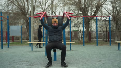 a coach wearing a brown beanie and black jacket is working out on outdoor equipment, lifting red bars over his head, in the background, a boy walks by, with passing cars in the distance