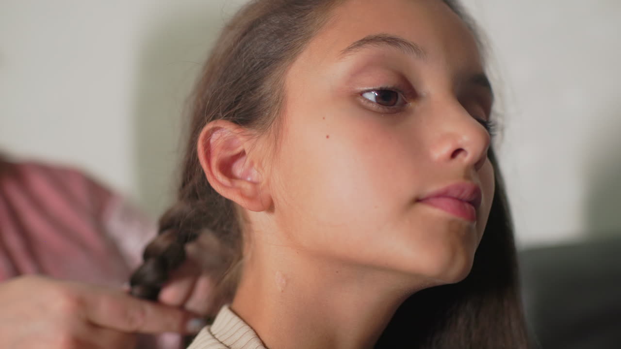 Nanny braiding girl’s hair, close-up shot of detailed grooming, child calmly sitting, intimate caregiving moment, peaceful indoor environment, personal touch, gentle, motherly bond