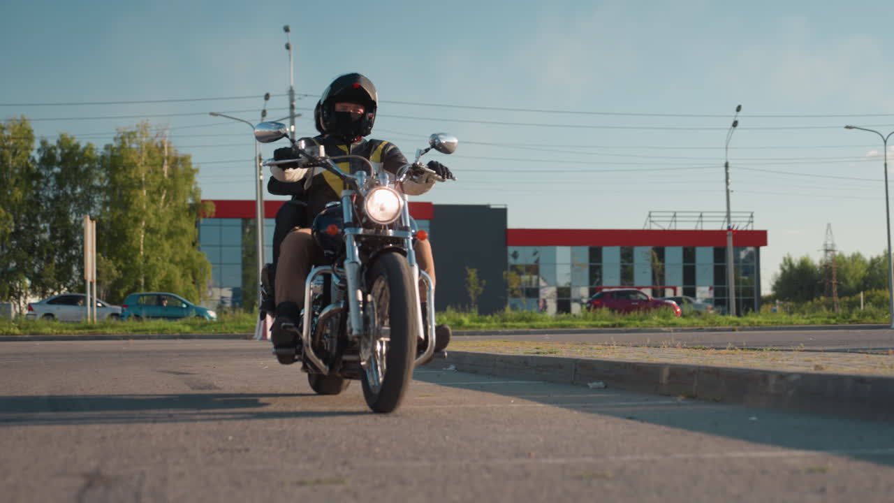 Motorcycle rider wearing helmet and leather jacket drives off with passenger seated behind under bright sunlight along urban road with cars and buildings in background