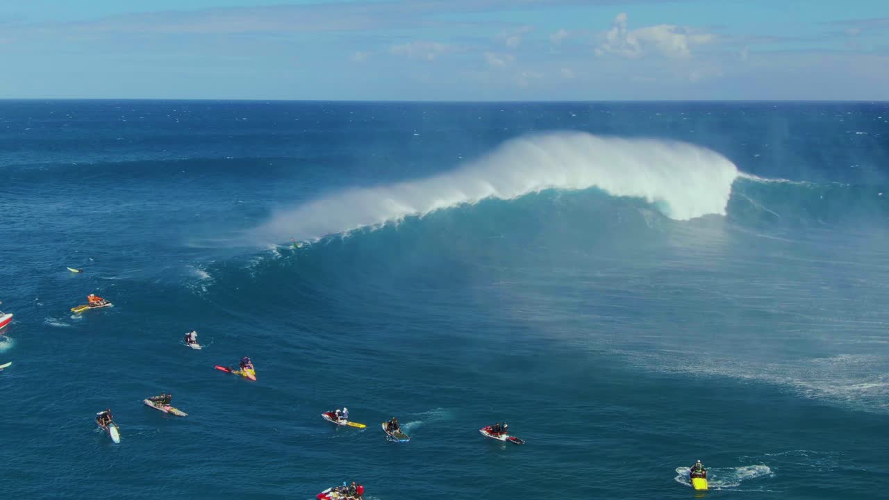 Big ocean wave spreading droplets around blue surface where surfers paddling followed by rescue boats in Hawaii