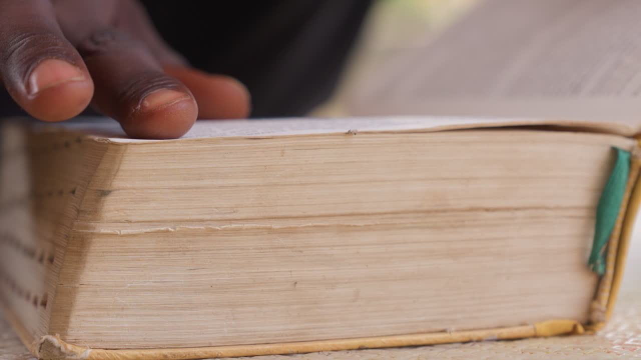 Pagine di un libro logoro sfogliate a mano, primo piano del dorso testurizzato e del segnalibro a nastro verde, movimento lento e deliberato, luce calda e soffusa, atmosfera di studio tranquilla, grana della carta visibile e margini invecchiati.