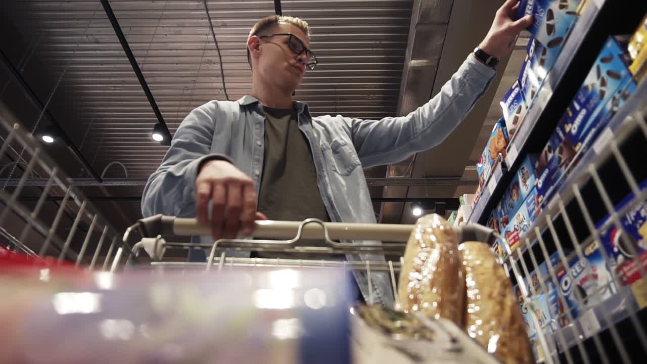 Male shopping. A man in glasses putting goods in shopping trolley. Passing through the dry goods aisle of a big supermarket. Low