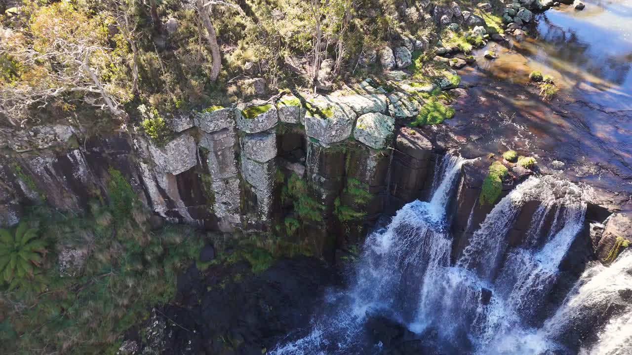Drone footage captures Ebor Falls cascading over basalt cliffs surrounded by lush forest in bright daylight, with smooth aerial camera movement and vibrant natural colors