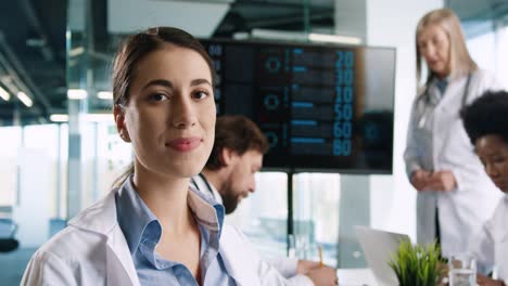 close-up view of young female doctor looking at camera and smiling while sitting at conference with mixed-races doctors discussing statistics of covid-19