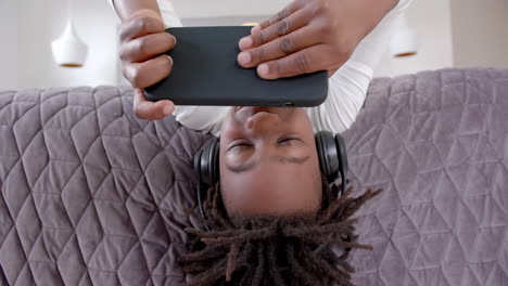 Happy-african-american-boy-wearing-headphones-lying-on-bed-using-smartphone-at-home,-slow-motion