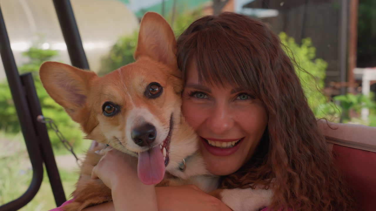 Smiling Woman Hugging Dog Outside, Happy Person Cuddling Her Corgi On Garden Swing In Summer Daylight, Caring Woman Shares Joyful Moment With Her Pet Dog Amidst Lush Garden Scenery In Sunlight