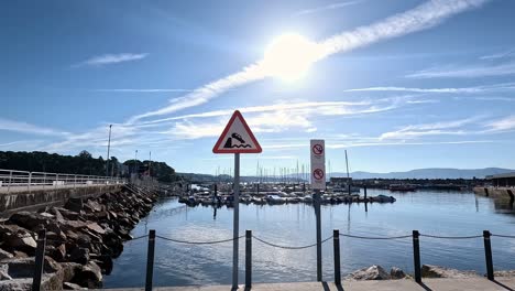 warning-signs-danger-of-falling-vehicles,-no-fishing-and-no-swimming-in-the-seaport-behind-chained-metal-poles,-sunlight-after-sunrise,-descriptive-panning