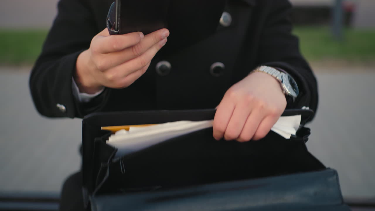 검은 옷을 입은 개인의 가까운 손이 가방에 서류를 검색하고 검은 일기를 가져오고, 초록색을 특징으로하는 배경, 불분명한 인물, 조용한 야외 환경에서 혀있는 경로